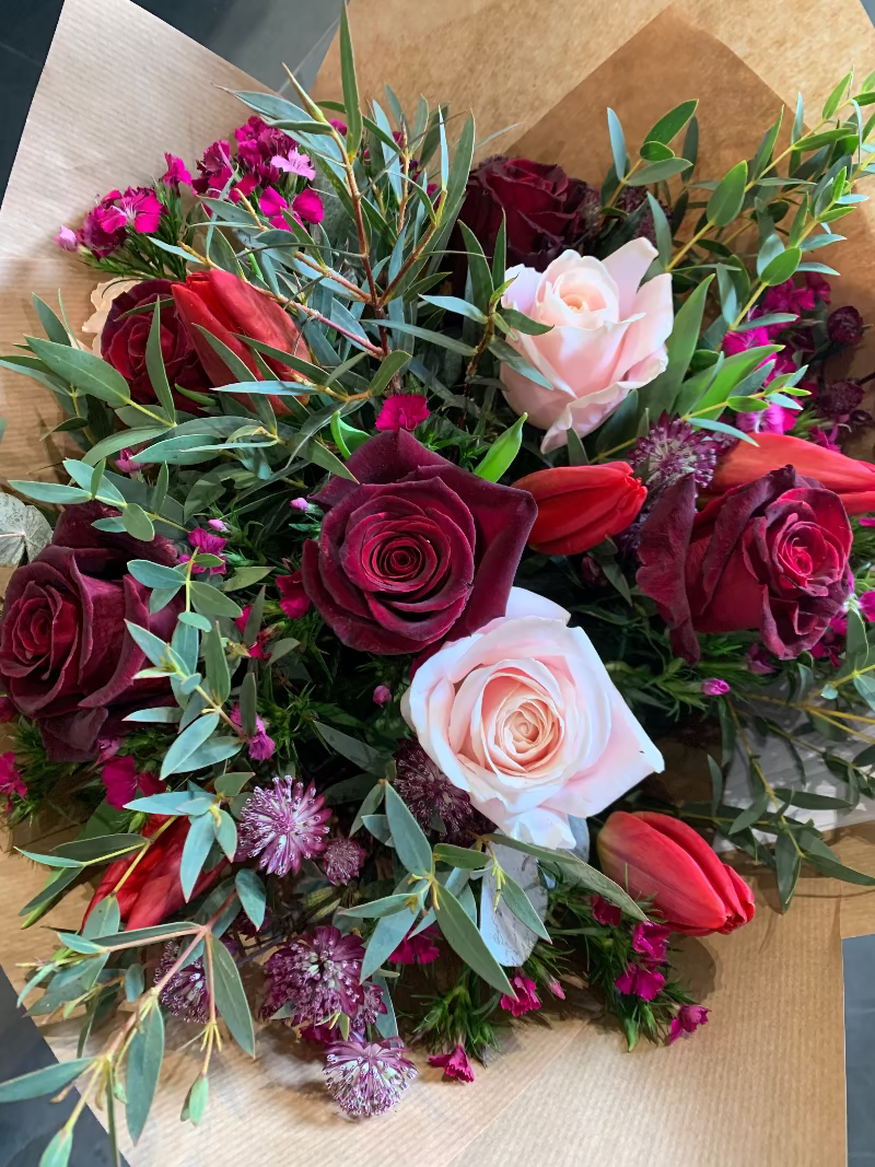 A romantic bouquet of red and pink flowers including roses and other seasonal foliage, wrapped in a paper with a velvet trim label and satin ribbon.