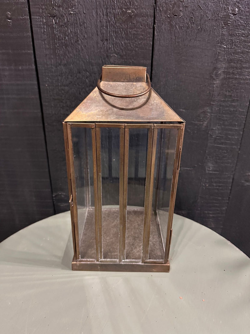 Antique brass finished glass panel lantern with slender glass panels and a visible aged appearance, placed against a wooden backdrop.