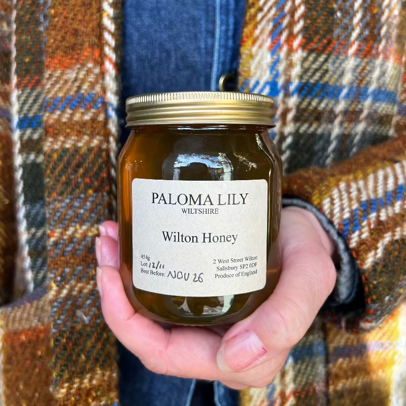 A hand holding a jar of Paloma Lily Wilton Honey, with the label clearly visible. The background features a person wearing a plaid scarf.