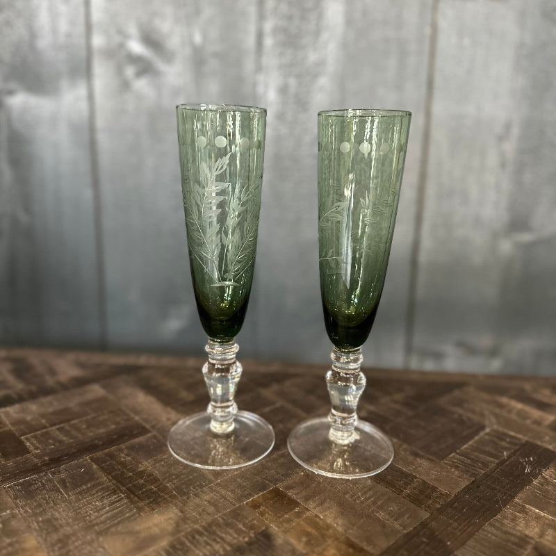 Two green etched champagne flutes on a wooden surface with a gray background